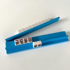 Puede incluir: Un estante azul para fichas de Mahjong con fichas blancas y rojas. El estante est&aacute; hecho de pl&aacute;stico azul brillante y sujeta las fichas en su lugar. Las fichas tienen dise&ntilde;os rojos y verdes y caracteres chinos.