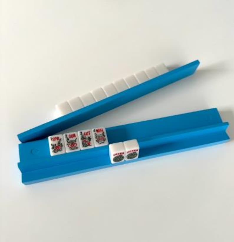 Puede incluir: Un estante azul para fichas de Mahjong con fichas blancas y rojas. El estante est&aacute; hecho de pl&aacute;stico azul brillante y sujeta las fichas en su lugar. Las fichas tienen dise&ntilde;os rojos y verdes y caracteres chinos.