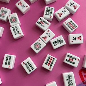 May include: A collection of white Mahjong tiles scattered across a pink surface. Each rectangular tile features engraved symbols, Chinese characters, and numbers in red, green, and black ink, including dragon motifs and various numerical suit patterns.