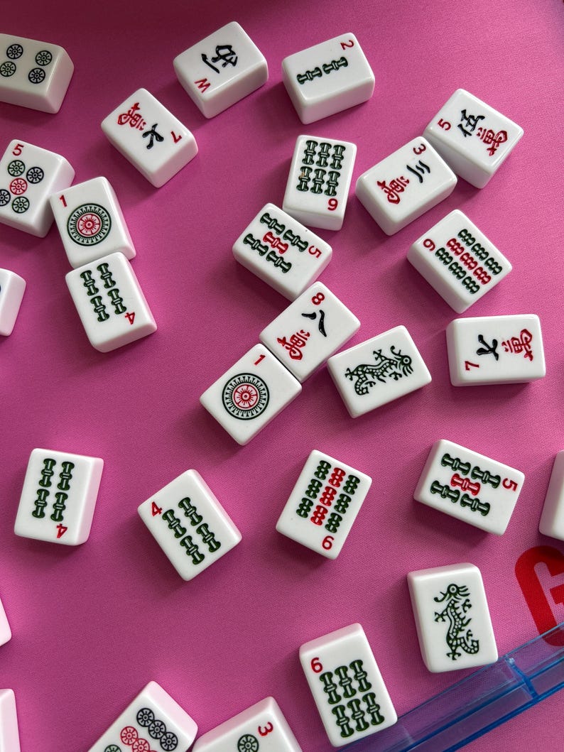 May include: A collection of white Mahjong tiles scattered across a pink surface. Each rectangular tile features engraved symbols, Chinese characters, and numbers in red, green, and black ink, including dragon motifs and various numerical suit patterns.