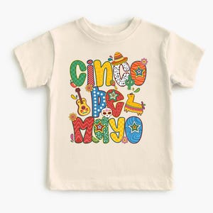 May include: A cream-colored t-shirt featuring the words "Cinco de Mayo" in colorful, festive lettering. The design includes a sombrero, guitar, cactus, and a piñata. The shirt is a children's size.