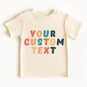 May include: Cream-colored t-shirt with the text "YOUR CUSTOM TEXT" in colorful, rounded letters. The text is in orange, teal, blue, red, yellow, and navy. The shirt has short sleeves and a crew neck.