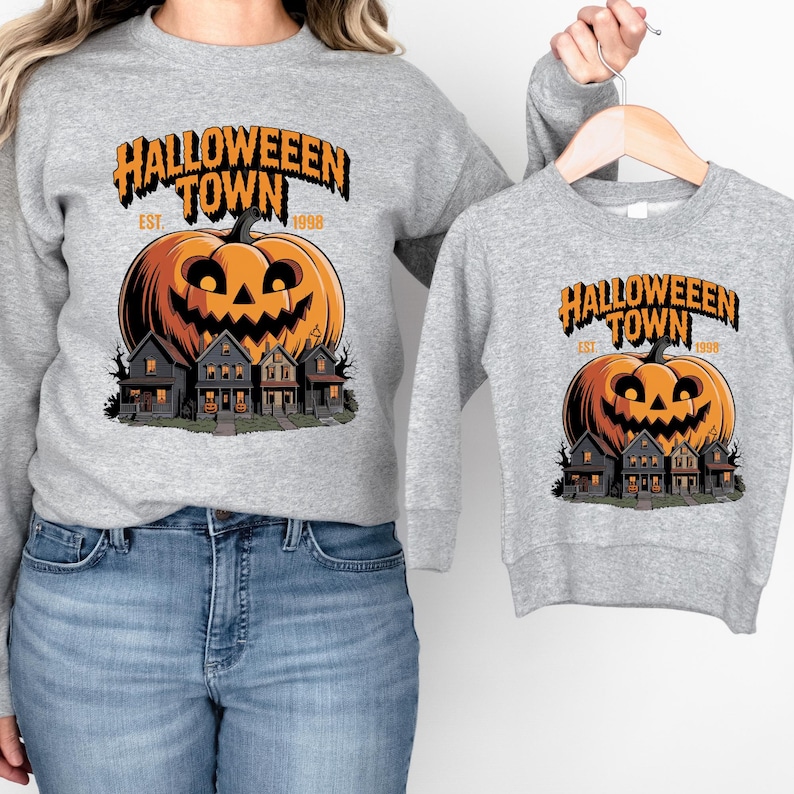 A woman wearing a gray sweatshirt with a Halloween-themed graphic of a pumpkin and houses, and a child wearing a smaller gray sweatshirt with the same Halloween-themed graphic.