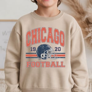 A young boy wearing a beige sweatshirt with a graphic design featuring a football helmet and the text "CHICAGO FOOTBALL" printed on it.