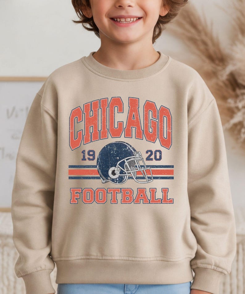A young boy wearing a beige sweatshirt with a graphic design featuring a football helmet and the text "CHICAGO FOOTBALL" printed on it.