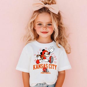 A young girl with blonde hair wearing a white t-shirt with a cartoon character and text printed on it.