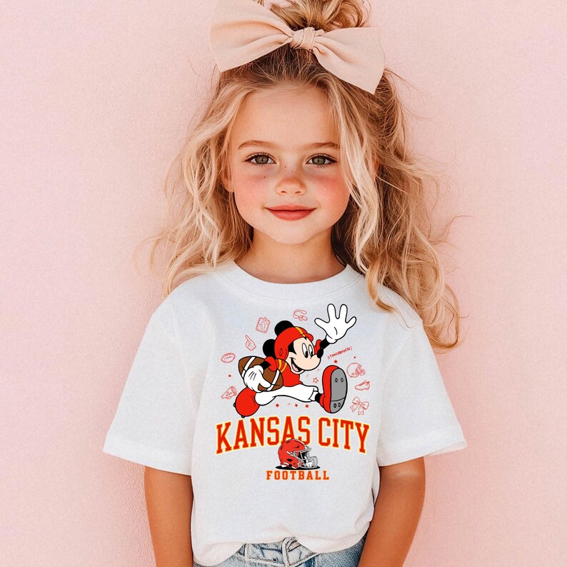 A young girl with blonde hair wearing a white t-shirt with a cartoon character and text printed on it.