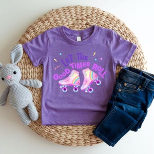 A purple t-shirt with a graphic of roller skates and the text "Let the good times roll" is displayed on a woven placemat, accompanied by a gray stuffed rabbit and a pair of blue jeans.