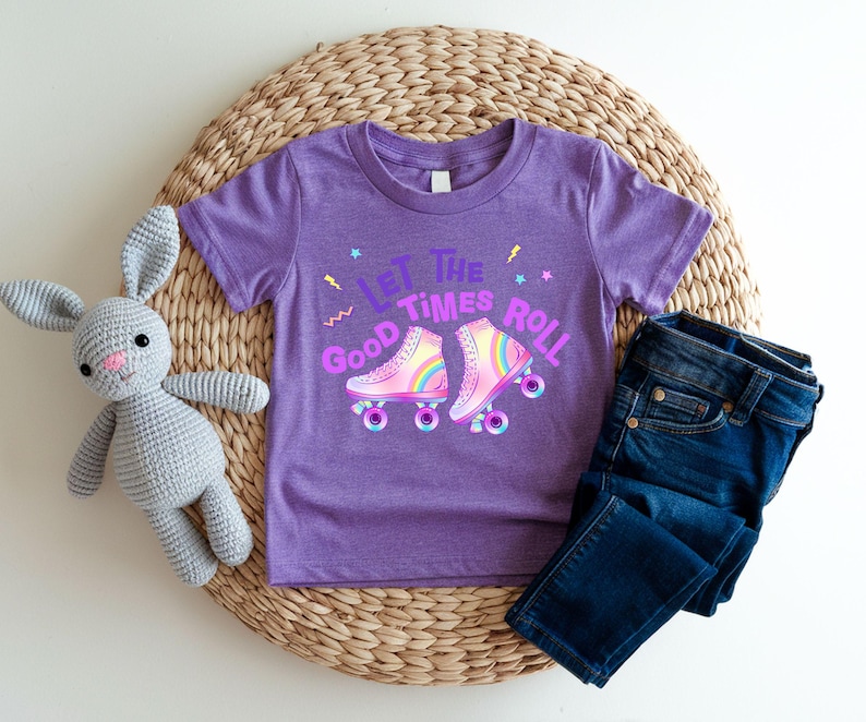 A purple t-shirt with a graphic of roller skates and the text "Let the good times roll" is displayed on a woven placemat, accompanied by a gray stuffed rabbit and a pair of blue jeans.
