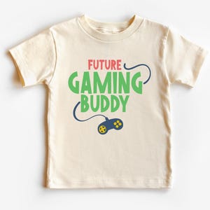 May include: A cream-colored short-sleeve t-shirt featuring the text FUTURE GAMING BUDDY in red and green lettering. Below the text is a dark blue video game controller graphic with yellow buttons and a cord that curves around the words.