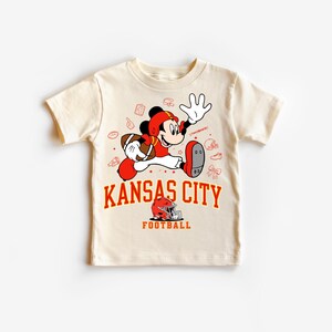 A white t-shirt with a cartoon image of Mickey Mouse holding a football and the text "KANSAS CITY FOOTBALL" printed on it.