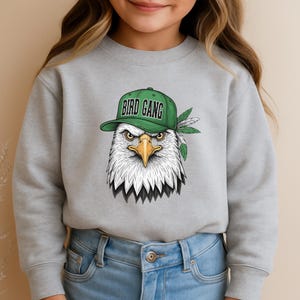 A young girl wearing a gray sweatshirt with a graphic of a bald eagle wearing a green baseball cap with the text "BIRD GANG" printed on it.