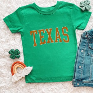 a green shirt with the word texas on it next to a pair of jeans and
