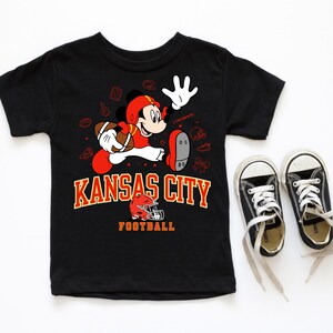 A black t-shirt with a graphic of Mickey Mouse and the text "KANSAS CITY FOOTBALL" printed on it, along with a pair of black and white sneakers.