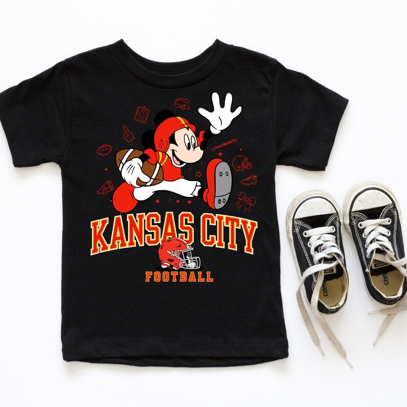 A black t-shirt with a graphic of Mickey Mouse and the text "KANSAS CITY FOOTBALL" printed on it, along with a pair of black and white sneakers.