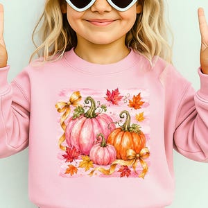 May include: A light pink crewneck sweatshirt featuring a graphic print of three pumpkins in shades of pink and orange, accented with gold ribbons and scattered autumn maple leaves against a soft pink brushstroke background.