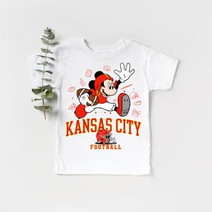 A white t-shirt with a graphic design featuring a cartoon character in a football uniform, along with the text "KANSAS CITY FOOTBALL".