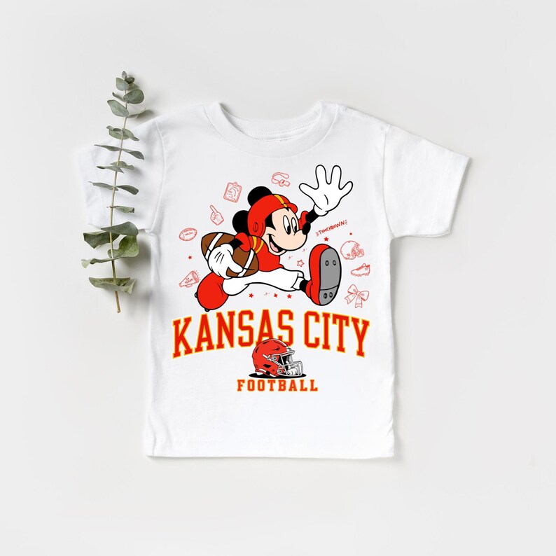 A white t-shirt with a graphic design featuring a cartoon character in a football uniform, along with the text "KANSAS CITY FOOTBALL".