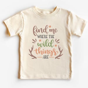 May include: A cream-colored short-sleeve t-shirt featuring the text "find me WHERE THE wild things ARE" in brown, green, and orange. The design includes small orange flower accents and two brown deer antler illustrations on the sides.