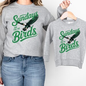 A woman wearing a gray sweatshirt with a graphic of an eagle and the text "Sunday are for the Birds" is holding a smaller gray sweatshirt with the same graphic and text.
