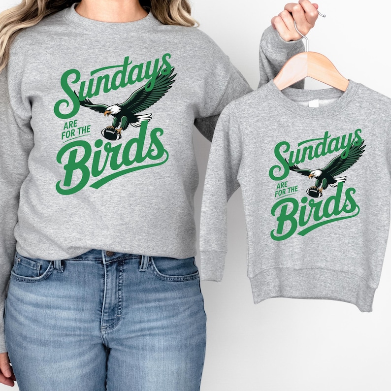 A woman wearing a gray sweatshirt with a graphic of an eagle and the text "Sunday are for the Birds" is holding a smaller gray sweatshirt with the same graphic and text.
