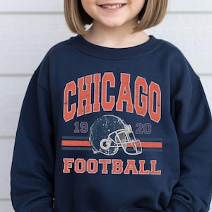 A young girl wearing a navy blue sweatshirt with a graphic of a football helmet and the text "CHICAGO FOOTBALL" printed on it.
