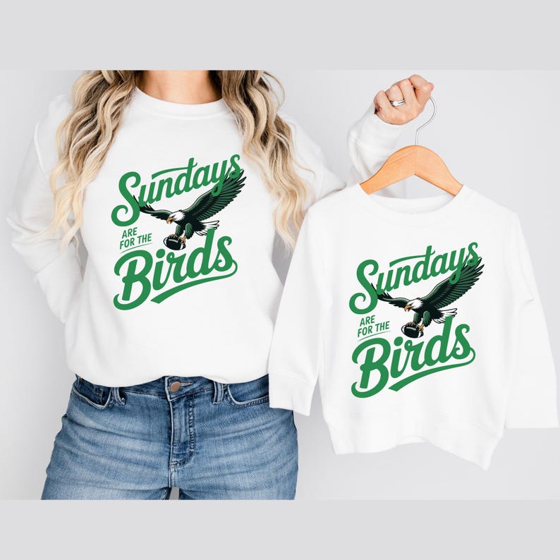 A woman wearing a white sweatshirt with a graphic design of a bird and the text "SUNDAYS ARE FOR THE BIRDS" is holding a smaller white sweatshirt with the same design.