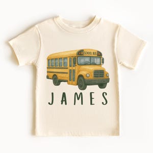 May include: A cream colored t-shirt with a yellow school bus illustration and the name "JAMES" printed in green.