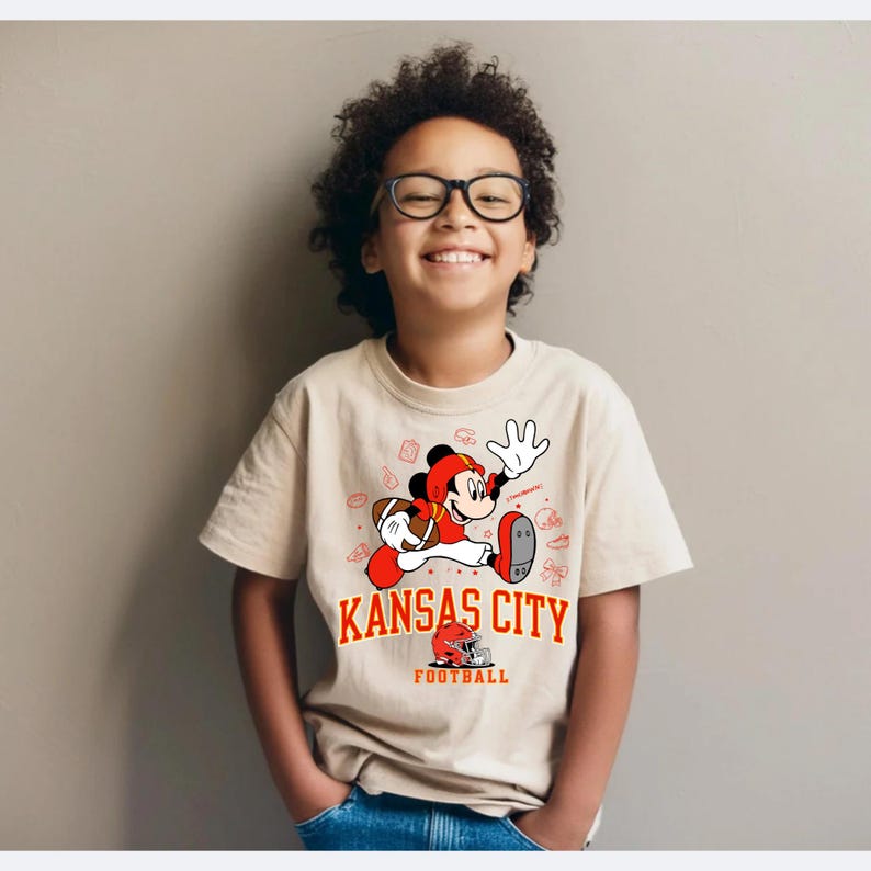 A young boy wearing glasses and a Kansas City football t-shirt is smiling and posing for the camera.