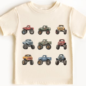May include: A cream-colored t-shirt with a graphic of nine different monster trucks in various colors, including red, blue, green, and yellow.