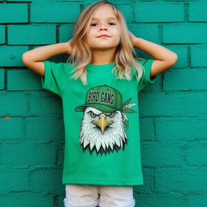 A young girl wearing a green t-shirt with a graphic of an eagle wearing a hat and the text "BIRD GANG" is standing in front of a green brick wall.