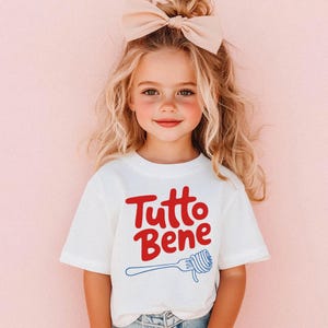 A young girl with blonde hair wearing a white t-shirt with the text "Tutto Bene" and a bow in her hair.