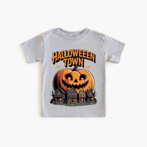 A gray t-shirt with a Halloween-themed graphic design. The design features a large jack-o-lantern and a spooky village scene.