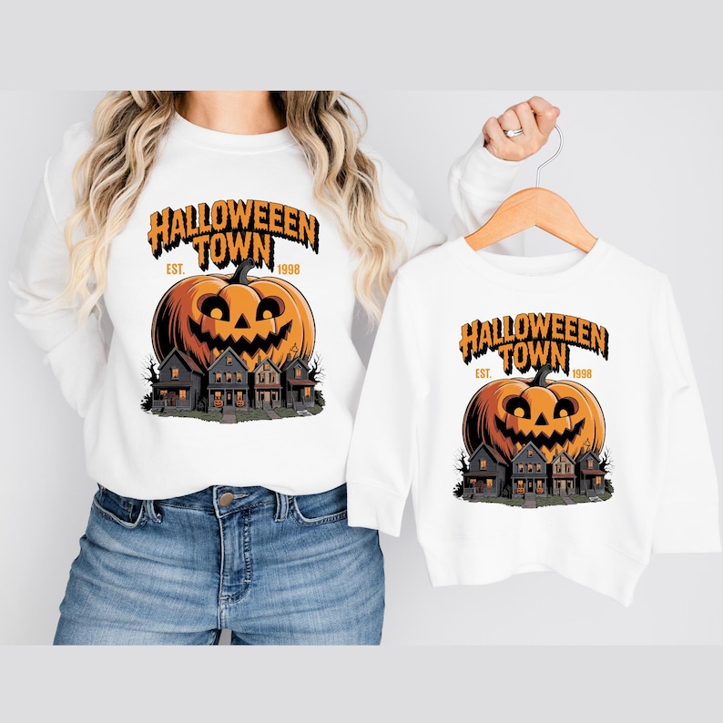 A woman wearing a white sweatshirt with a Halloween-themed graphic and a smaller white sweatshirt with a similar Halloween graphic.
