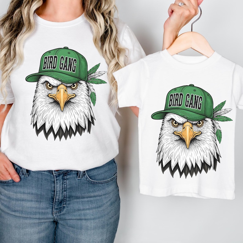 A woman wearing a white t-shirt with a graphic of an eagle wearing a green baseball cap and the text "BIRD GANG" printed on it, holding the t-shirt to the side.