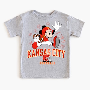 A gray t-shirt with a graphic design featuring a cartoon character in a red and white uniform, holding a football, and surrounded by various football-related icons and text.