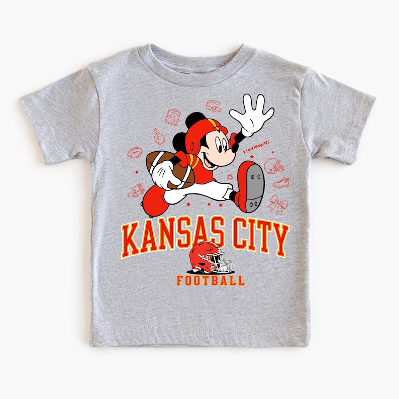 A gray t-shirt with a graphic design featuring a cartoon character in a red and white uniform, holding a football, and surrounded by various football-related icons and text.