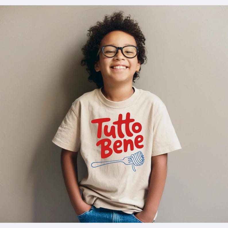 A young boy with curly hair and glasses is smiling and wearing a white t-shirt with the text "Totto Bene" printed on it.