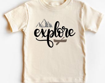 May include: A cream-colored short-sleeve t-shirt featuring a graphic print on the front. The design includes a gray mountain illustration above the black cursive text "explore" and a small brown canoe graphic positioned below the word.