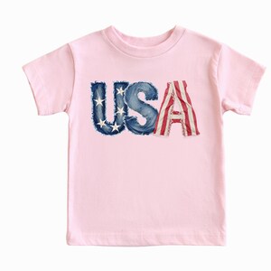 A pink t-shirt with the word "USA" printed on it, featuring the letters in a distressed, distressed-looking style.