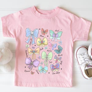 May include: Pink t-shirt with a colorful graphic of bows and the names of Disney princesses, including Ariel, Belle, Aurora, Tiana, Snow White, Merida, Mulan, Pocahontas, Rapunzel, Cinderella, Jasmine, and Moana.