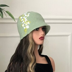 May include: A sage green crocheted cloche hat with a white and yellow floral design.