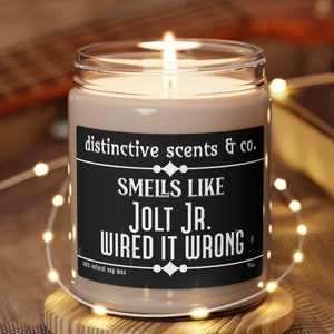 May include: A brown glass candle jar with a black label that reads "distinctive scents & co. SMELLS LIKE JOLT JR. WIRED IT WRONG 100% natural soy wax 9oz".