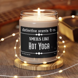 May include: A brown glass candle jar with a black label that reads "distinctive scents & co. Smells Like Hot Yoga" and "100% natural soy wax". The candle is lit and surrounded by white string lights.