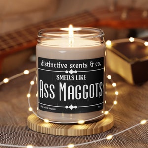 May include: A brown glass candle with a black label that reads "distinctive scents & co. Smells like Ass Maggots 100% natural soy wax 9oz".