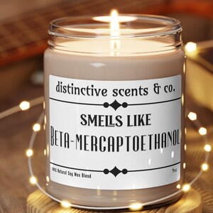 May include: A brown glass candle with a white label that reads "distinctive scents & co. SMEILS LIKE BETA-MERCAPTOETHANOL 100% Natural Soy Wax Blend 9oz".