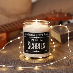 May include: A black candle jar with a white label that reads "distinctive scents & co. Smells like Scabies 100% Natural Soy Wax Blend". The candle is lit and the jar is sitting on a wooden coaster.