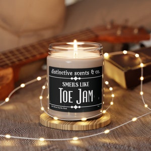 May include: A brown glass candle with a black label that reads "distinctive scents & co. Smells like Toe Jam." The candle is sitting on a wooden coaster and surrounded by white string lights.