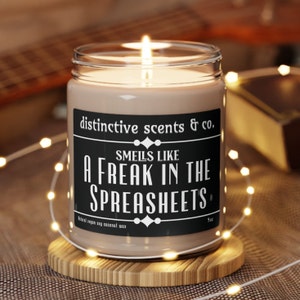 May include: A brown glass candle with a black label that reads "distinctive scents & co. Smells like a freak in the spreadsheets." The candle is lit and surrounded by white fairy lights.
