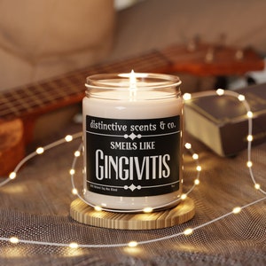 May include: A white candle in a glass jar with a black label that reads "distinctive scents & co. Smells like GINGIVITIS 100% Natural Soy Wax Blend". The candle is lit and sitting on a wooden coaster.